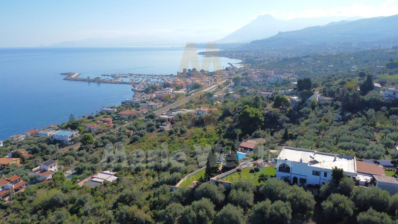 Aerial of the villa among olives, with the railway and coast road between the garden and the sea, and a marina breakwater below