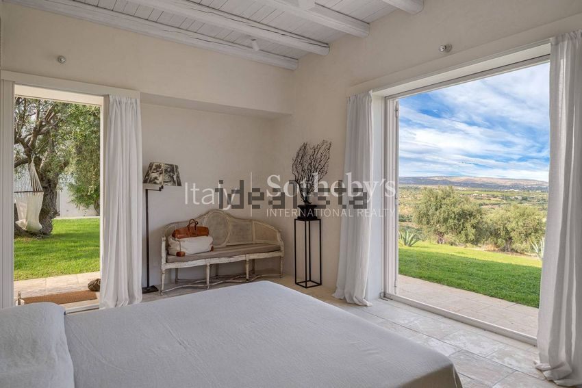 A bedroom with a bed, a bench, and open doors onto a lawn and distant hills