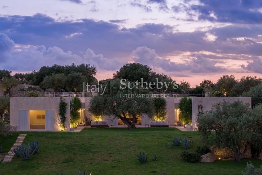The long one-storey front at dusk, lights on, an olive against the wall and agaves in the lawn