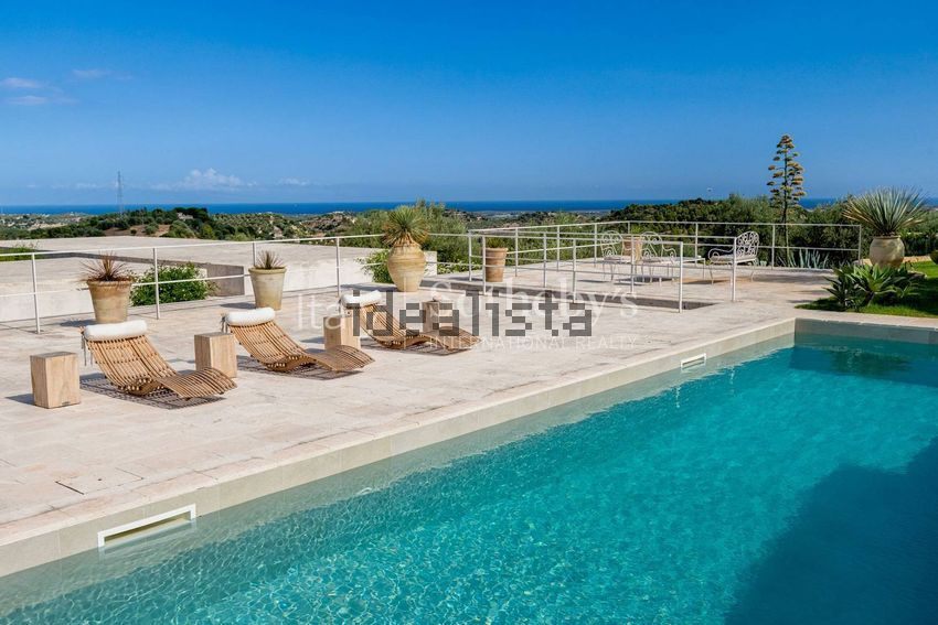 A turquoise pool with wooden loungers, and the sea on the horizon past a railing
