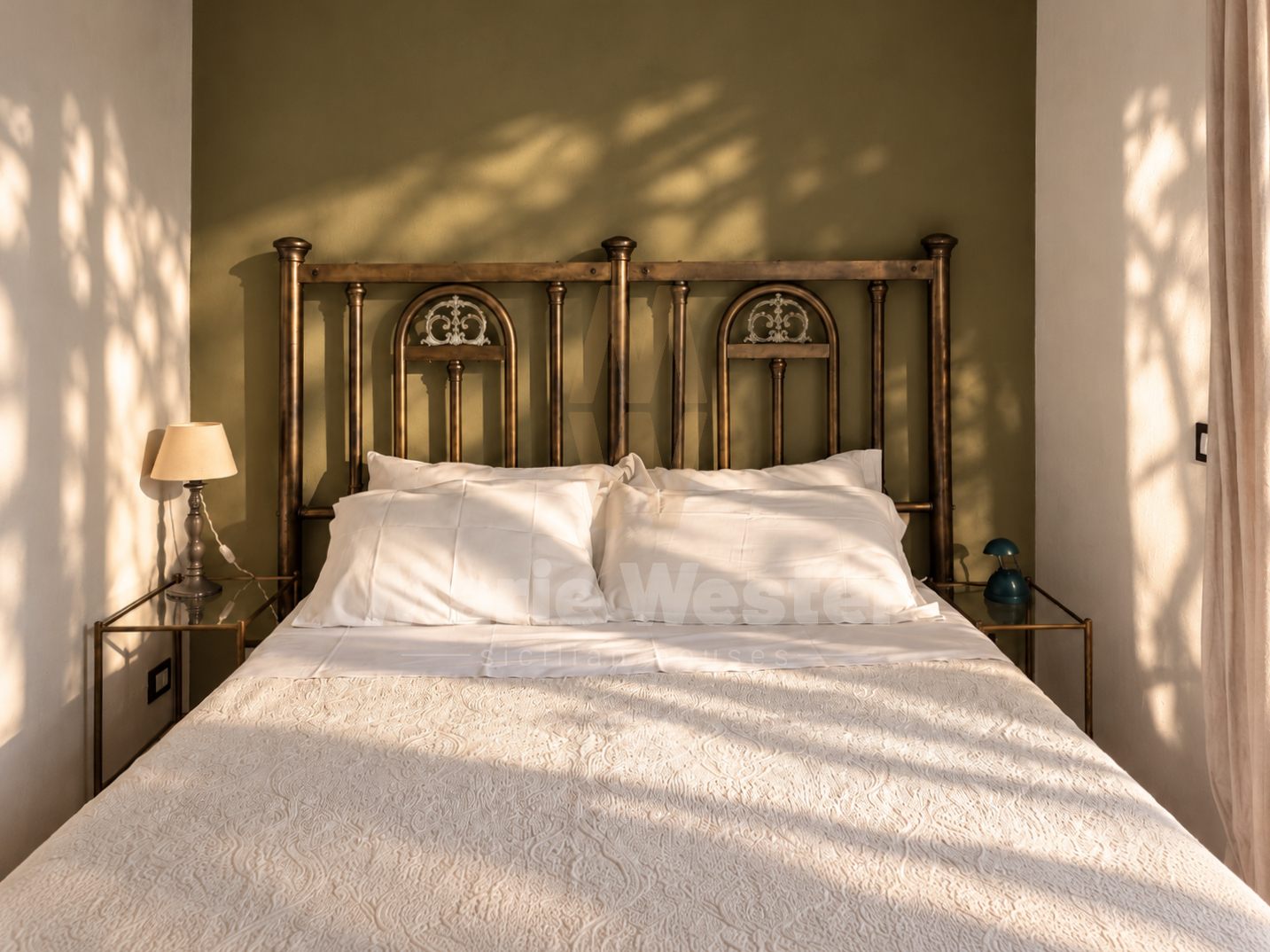 A bedroom with an olive-green wall and a brass bed in shuttered light