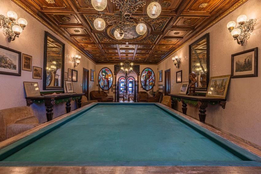 A billiard room under a coffered painted ceiling, with pictures, mirrors, and stained glass