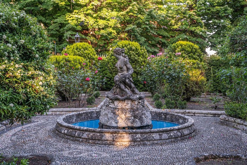 A stone figure seated on a shell in a round basin, on a pebble court among clipped bushes