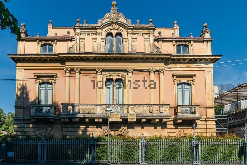 The ochre street front of the villa behind an iron fence, with paint loss on the upper storey and scaffolding at the right